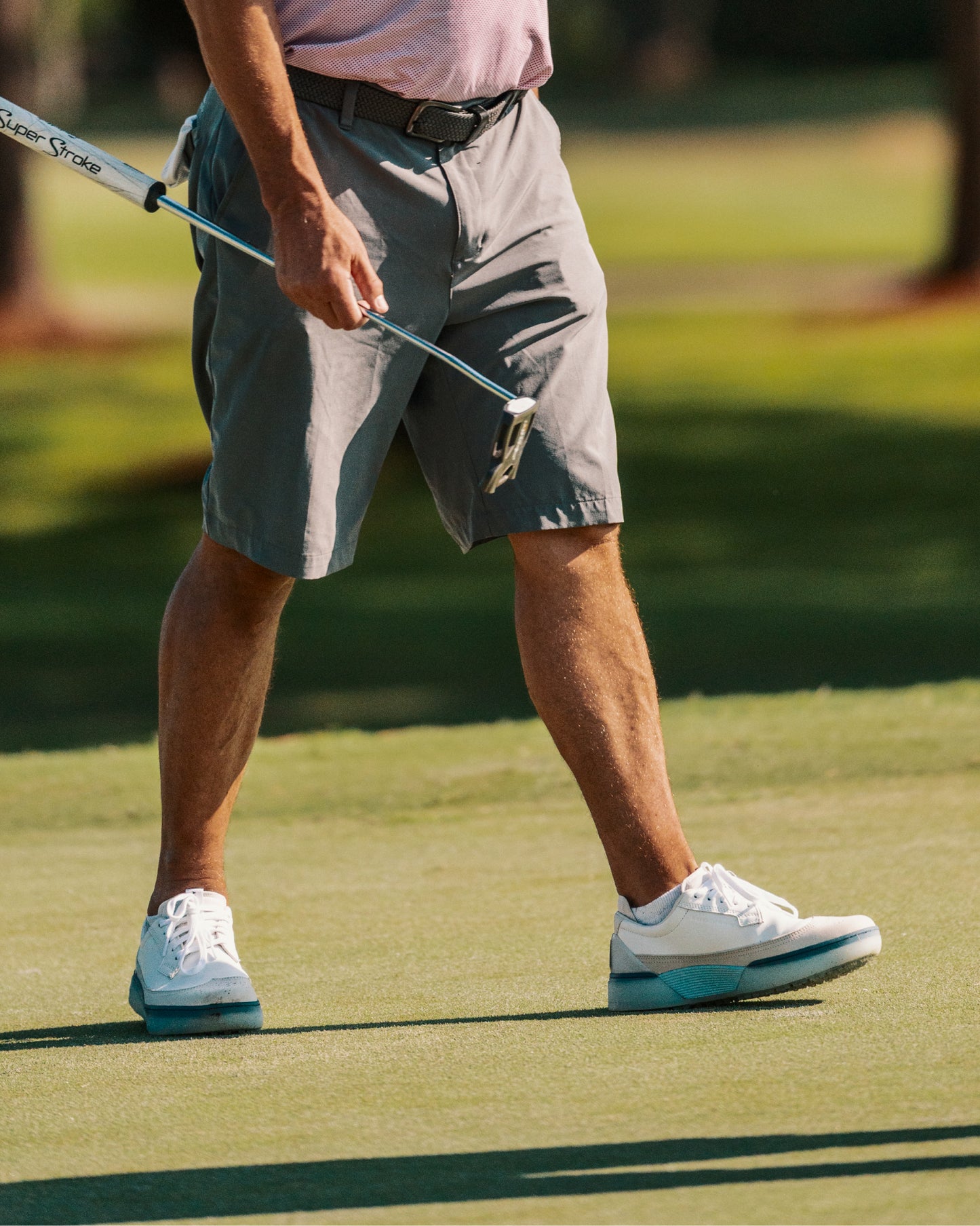 Golfer walking across a green with a putter, wearing white and aqua Beta Longwing golf shoes with midsole detail visible