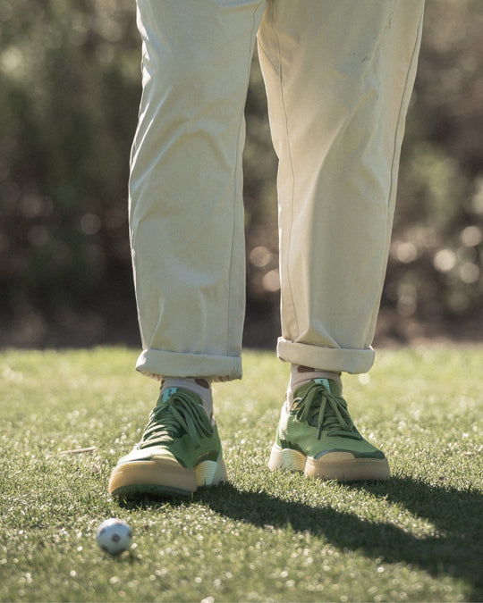 Person wearing Olive-Aqua Beta Lite golf shoes and beige pants standing on grass with a golf ball nearby.
