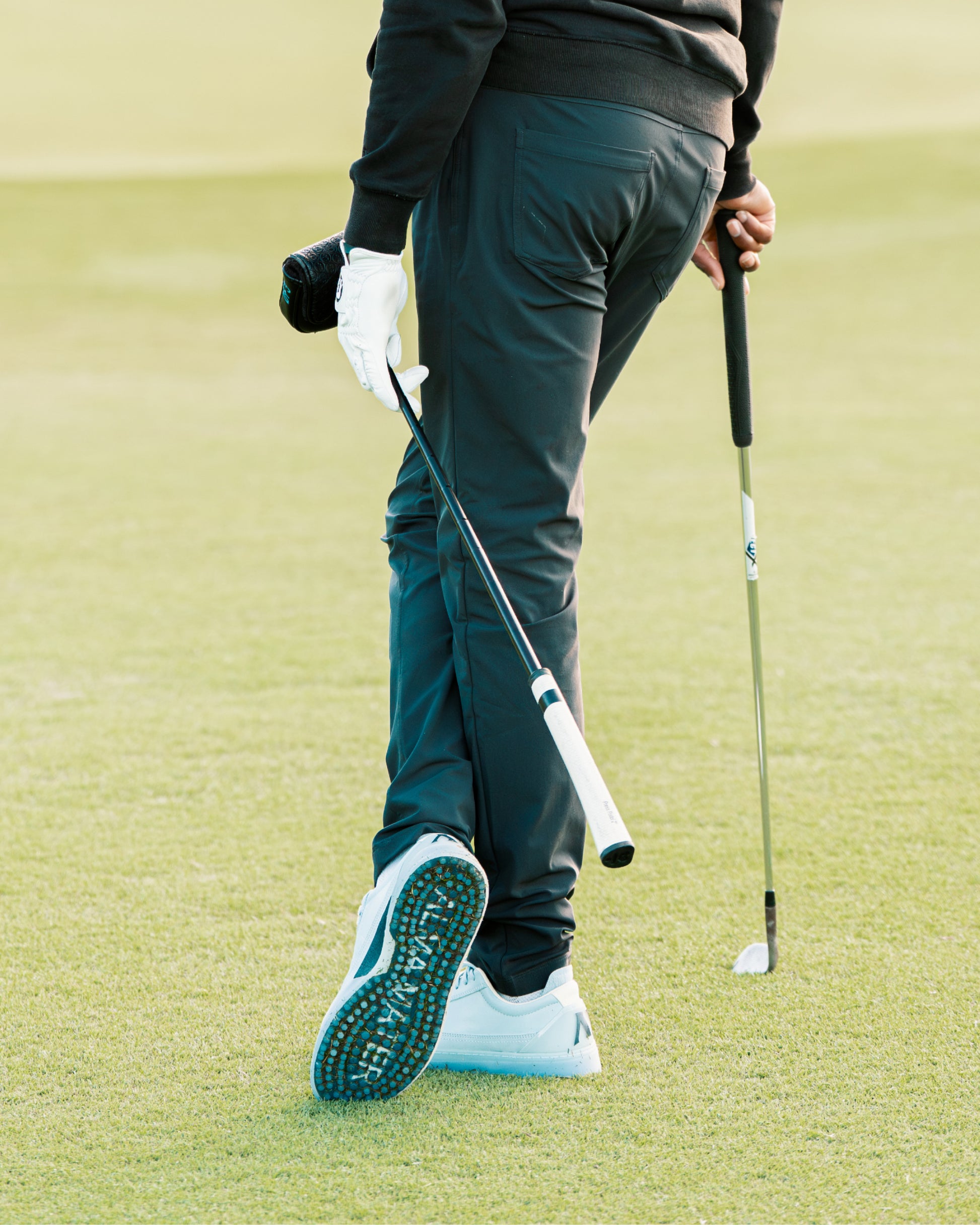 Golfer walking on a green holding clubs, wearing sky and navy Beta Longwing golf shoes with outsole visible