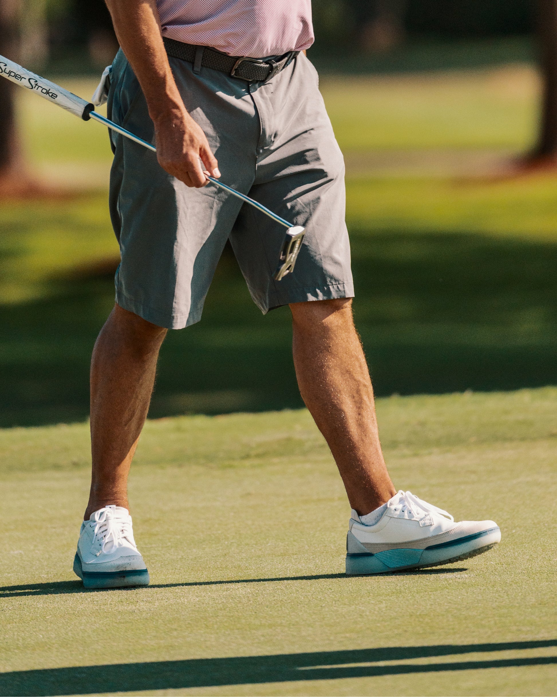 Golfer walking across a green with a putter, wearing white and aqua Beta Longwing golf shoes with midsole detail visible