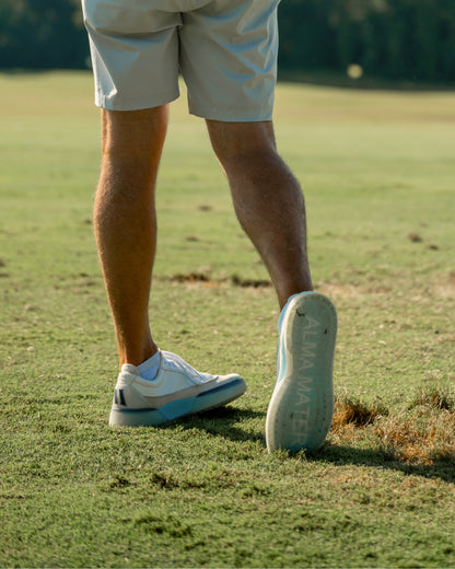 Golfer walking on grass with one foot lifted, showing the outsole of white and aqua Beta Longwing golf shoes