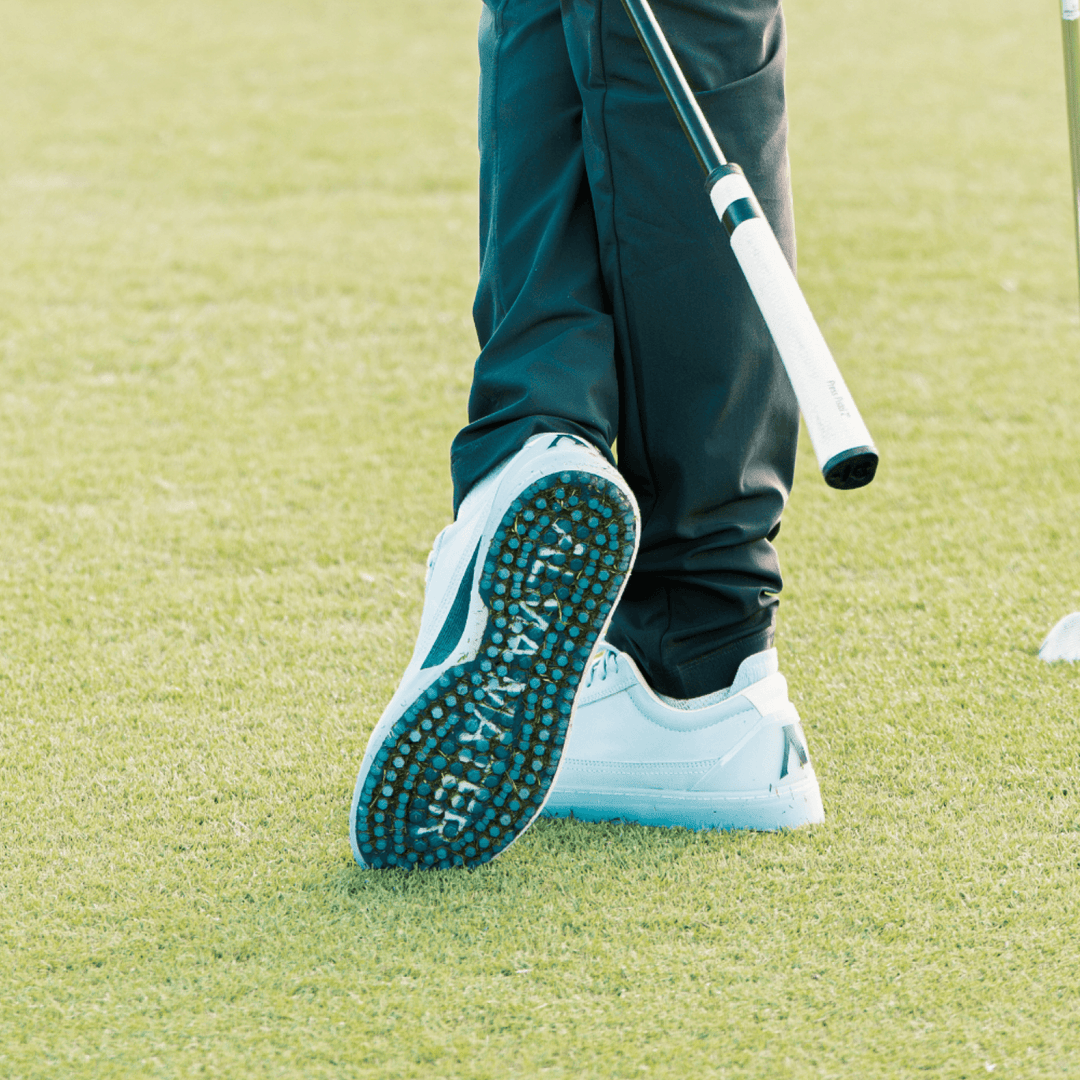 Golfer walking on fairway grass, heel lifted mid-step to reveal the Shark-Tooth™ traction outsole of a white and teal Alma Mater golf shoe, with ALMA MATER branding visible across the sole.