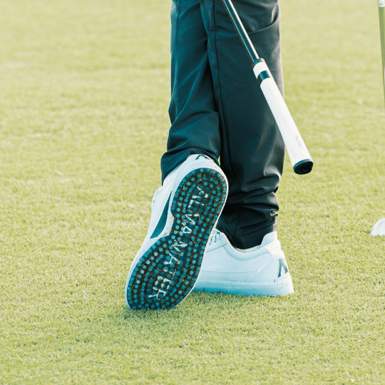 Golfer walking on fairway grass, heel lifted mid-step to reveal the Shark-Tooth™ traction outsole of a white and teal Alma Mater golf shoe, with ALMA MATER branding visible across the sole.