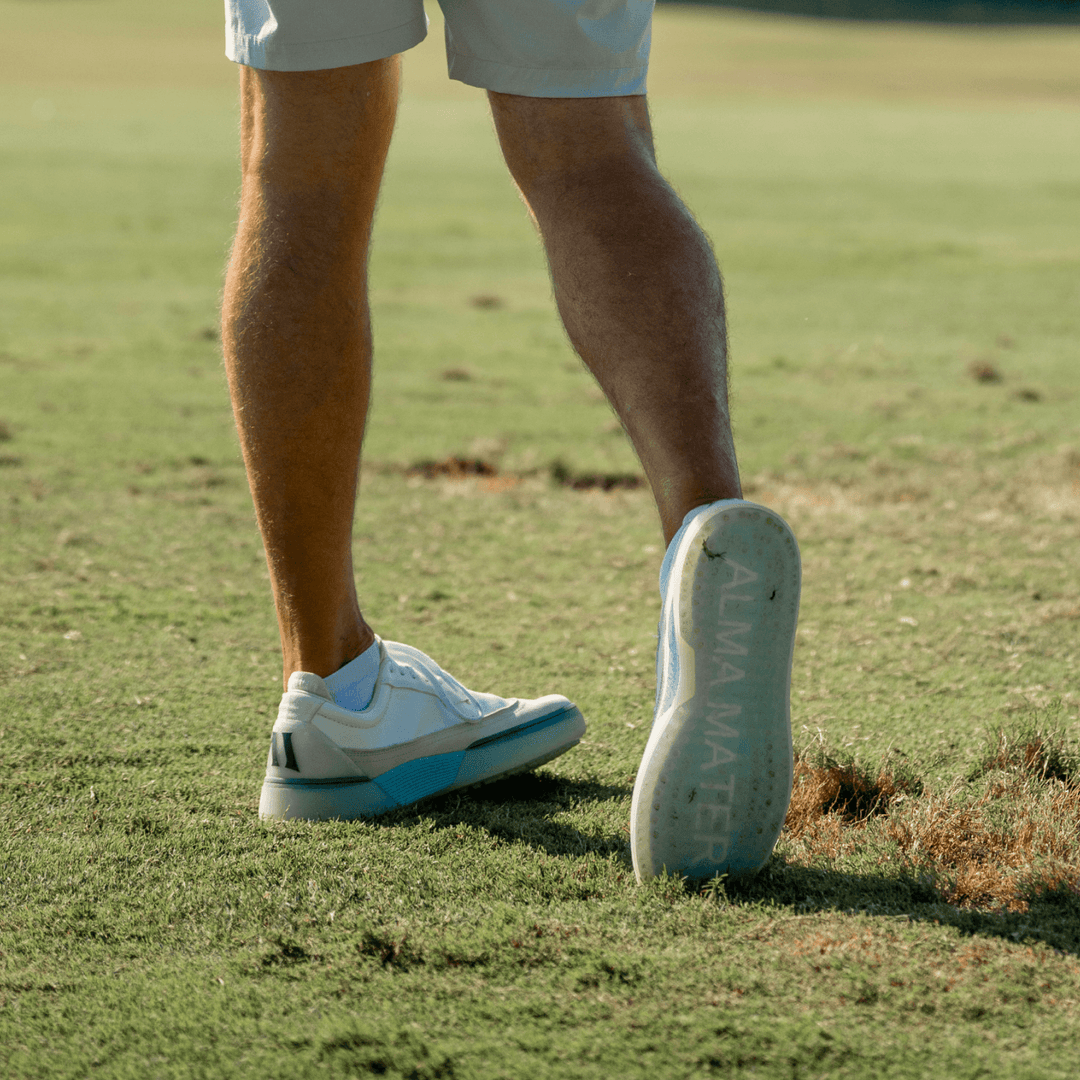 Golfer walking across the fairway in white and navy Beta Longwing golf shoes, heel lifted to reveal the translucent outsole with ALMA MATER branding, shown in warm late-afternoon light.