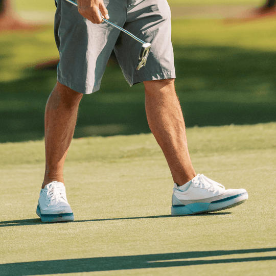 Golfer standing on a sunlit green holding a putter, wearing Beta Longwing white and navy Alma Mater golf shoes with a sculpted blue midsole and spikeless outsole, shown in real play on the course.