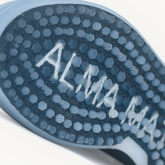 Close-up of a shoe sole with 'Alpina' branding on a white background. Highlighting the Shark-Tooth™ Activated outsole with visible traction pattern and ALMA MATER branding across the sole.