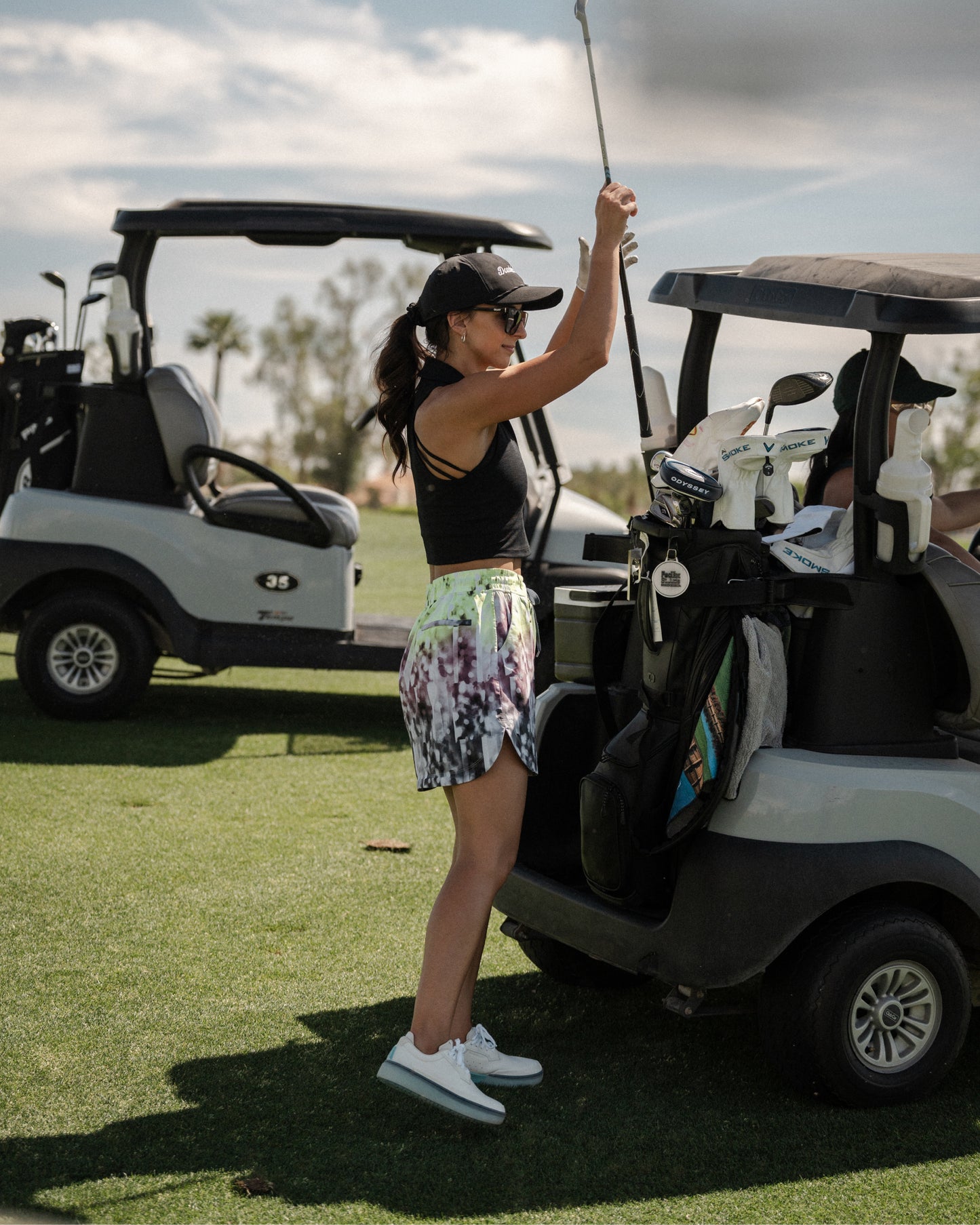 Woman standing beside a golf cart holding a club, wearing white and aqua Beta Derby golf shoes