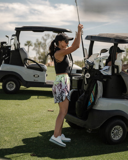 Woman standing beside a golf cart holding a club, wearing white and aqua Beta Derby golf shoes