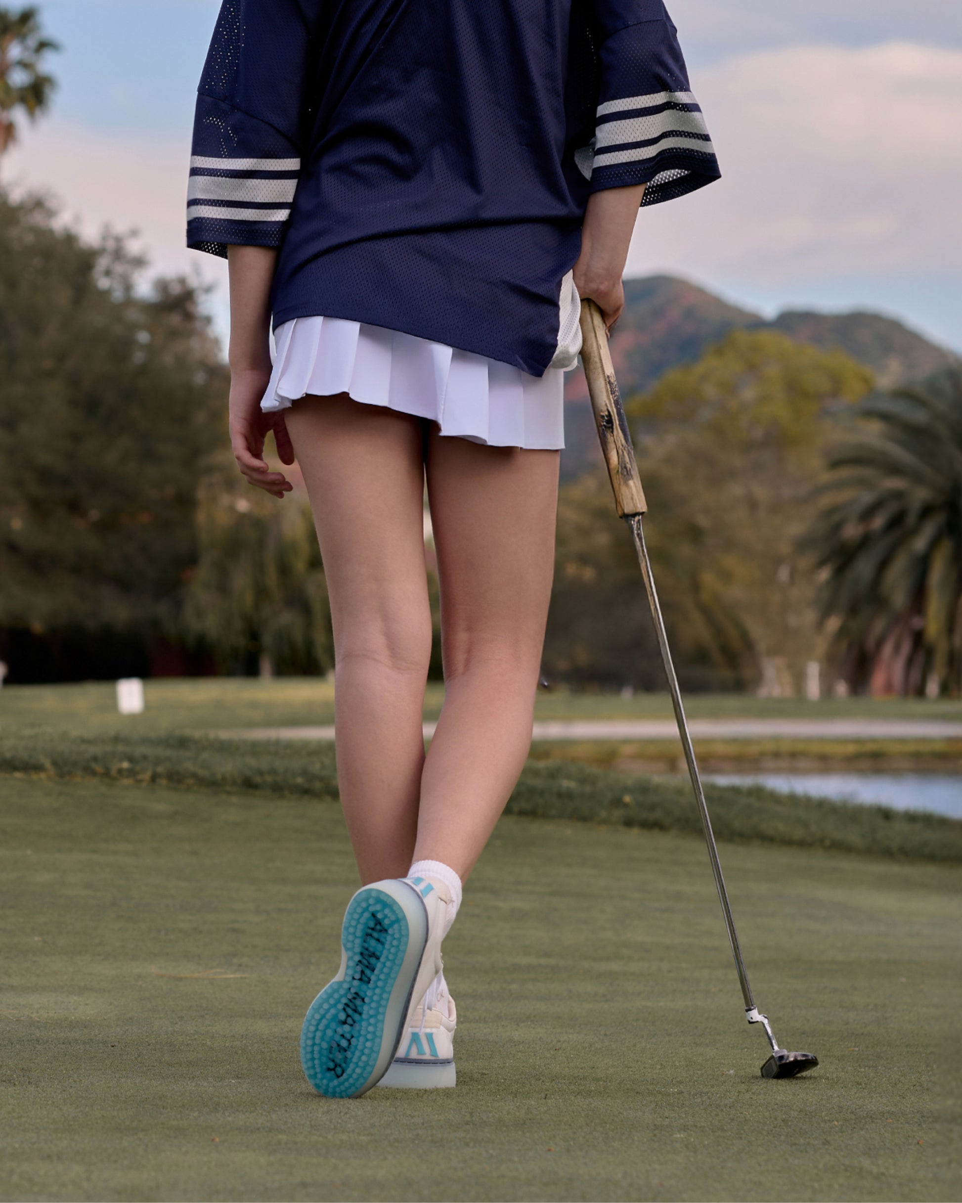 Woman walking on a golf green holding a putter, wearing white and aqua Beta Derby golf shoes with outsole visible