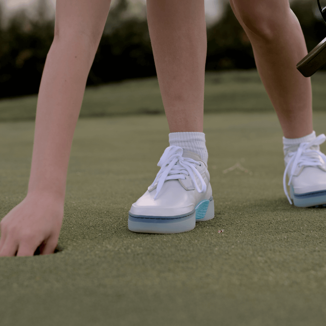 Close-up of women’s Beta Derby golf shoe in white and aqua as a golfer reaches to pick up a ball on the green.