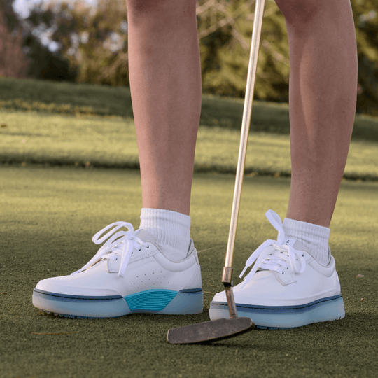 Women’s Beta Derby golf shoes in white and aqua on the putting green beside a putter, full-grain leather upper with spikeless outsole.