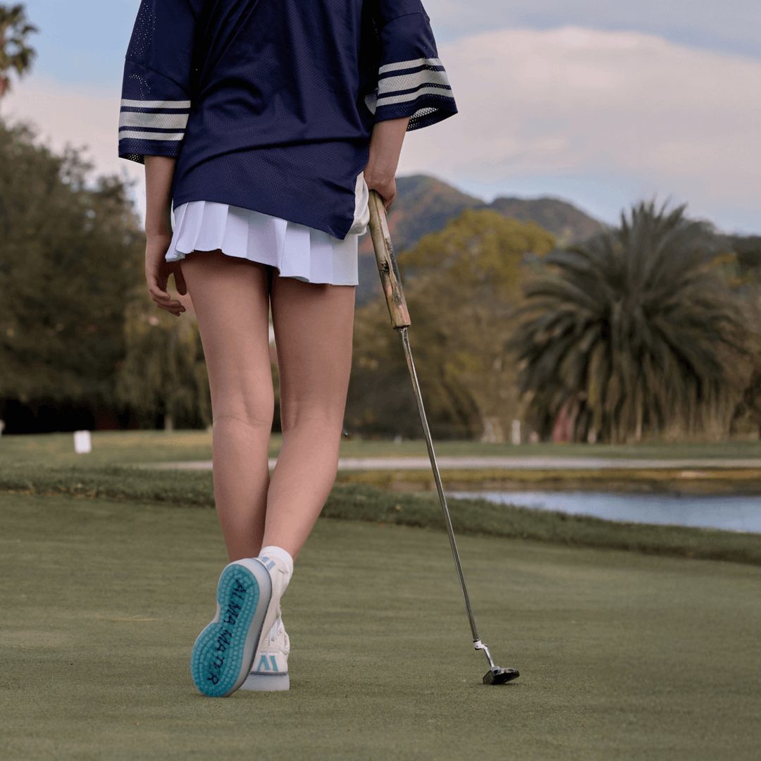 Rear view of women’s Beta Derby golf shoe in white and aqua walking across the green, showing aqua spikeless outsole traction.