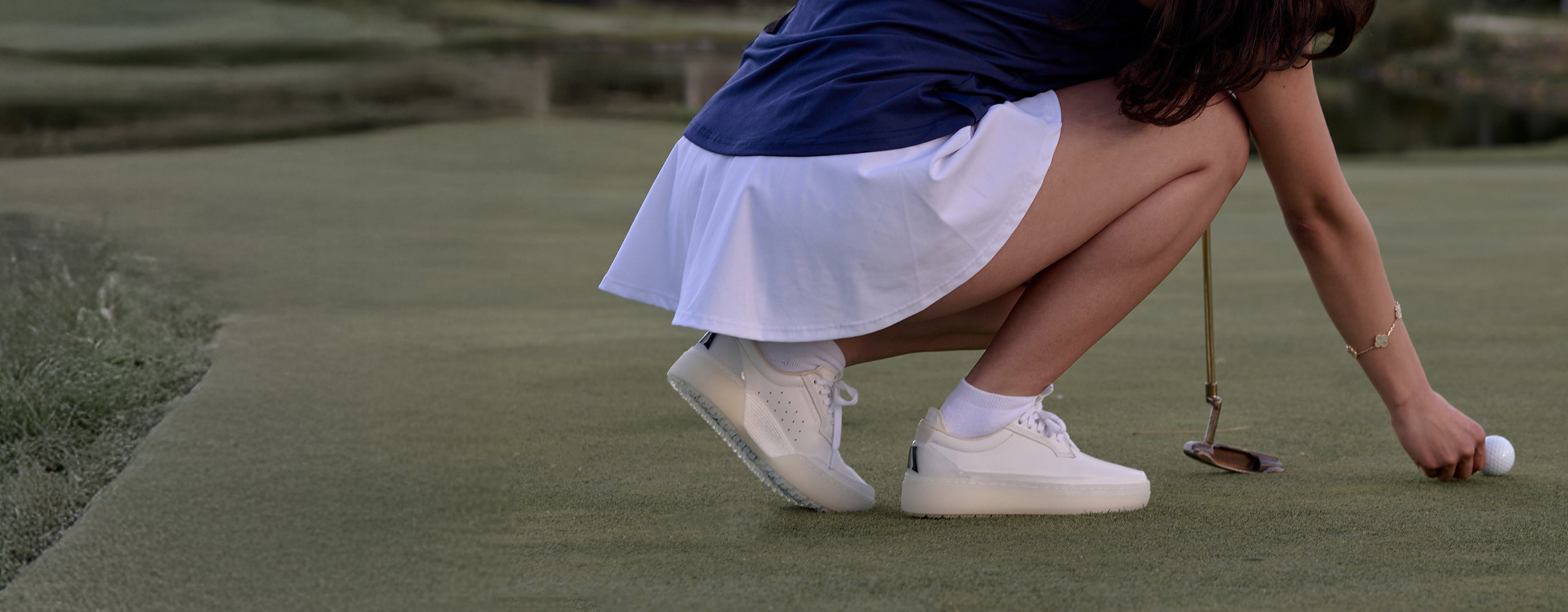 Woman in blue top and white skirt crouching on a golf green, placing a golf ball, wearing white Beta Derby sneakers
