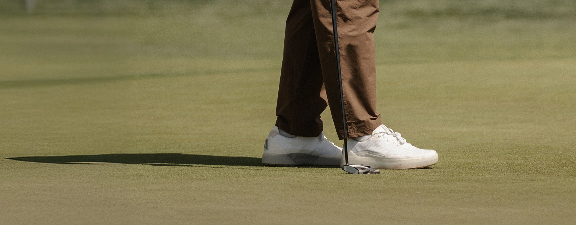Person on a golf course with white BEta Lite shoes and brown pants, holding a golf club.