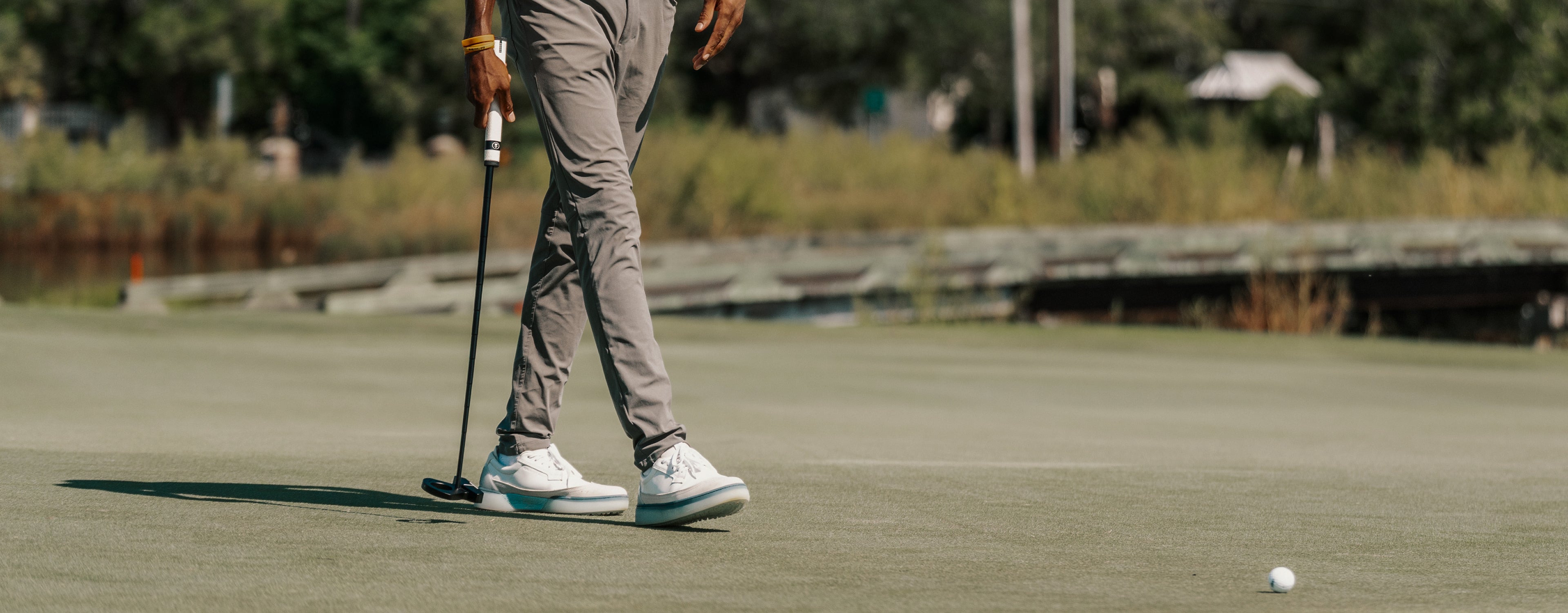 Person walking on a golf green holding a putter, wearing white and navy Beta Longwing golf shoes